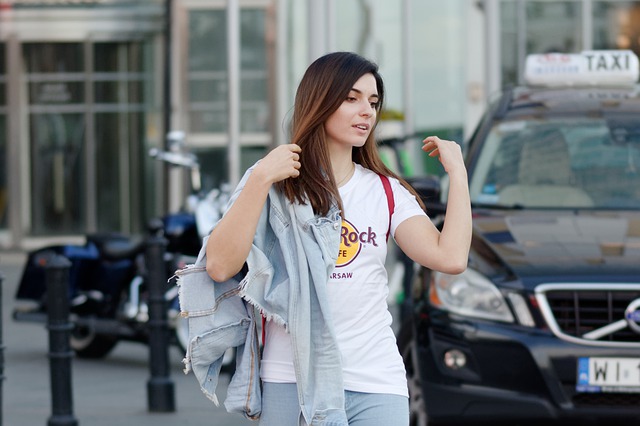 Žena v bielom tričku s nápisom a rifľovou nudnou v ruke ide po ulici.jpg