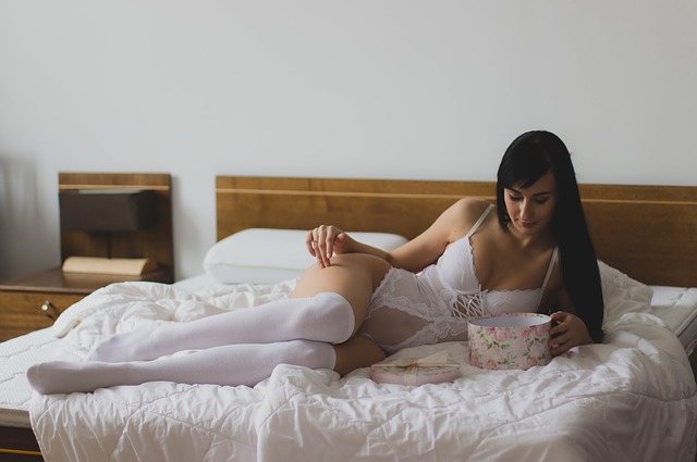 Žena v bielej erotickej bielizni leží na posteli s ružovým poťahom a pozerá do krabičky.jpg
